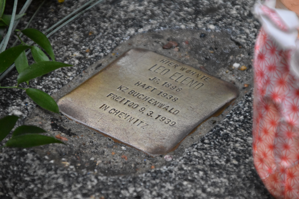 Das Bild zeigt den Stolperstein für Leo Elend umrahmt von einer Kerze im Glas und einer Blume.